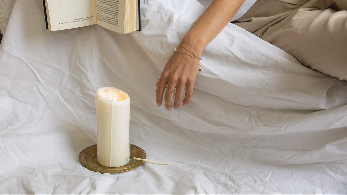 The LLIOR Signature Candle on a brass dish on a bed, beside someone reading